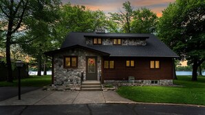 A two-story log cabin with a stone facade and a dark roof, surrounded by lush greenery and a lake view. - Dock, Firepit & Cuyuna Trails at Serpent Lake (Crosby)