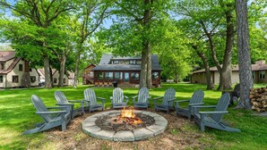 A fire pit surrounded by Adirondack chairs in a grassy area with a large house in the background. - Dock, Firepit & Cuyuna Trails at Serpent Lake (Crosby)