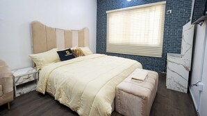 A bedroom with a bed, bedside tables, a nightstand, a bench, a window with blinds, and a wall-mounted air conditioning unit. - Idiroko estate - Green, Beautiful, Quiet and Central (Lagos)