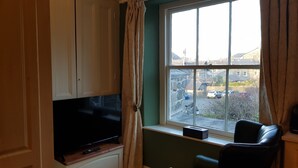 A living room with a television on a wooden cabinet, a black leather chair, and a window with a view of a street and buildings. - Afallon Townhouse (Dolgellau)