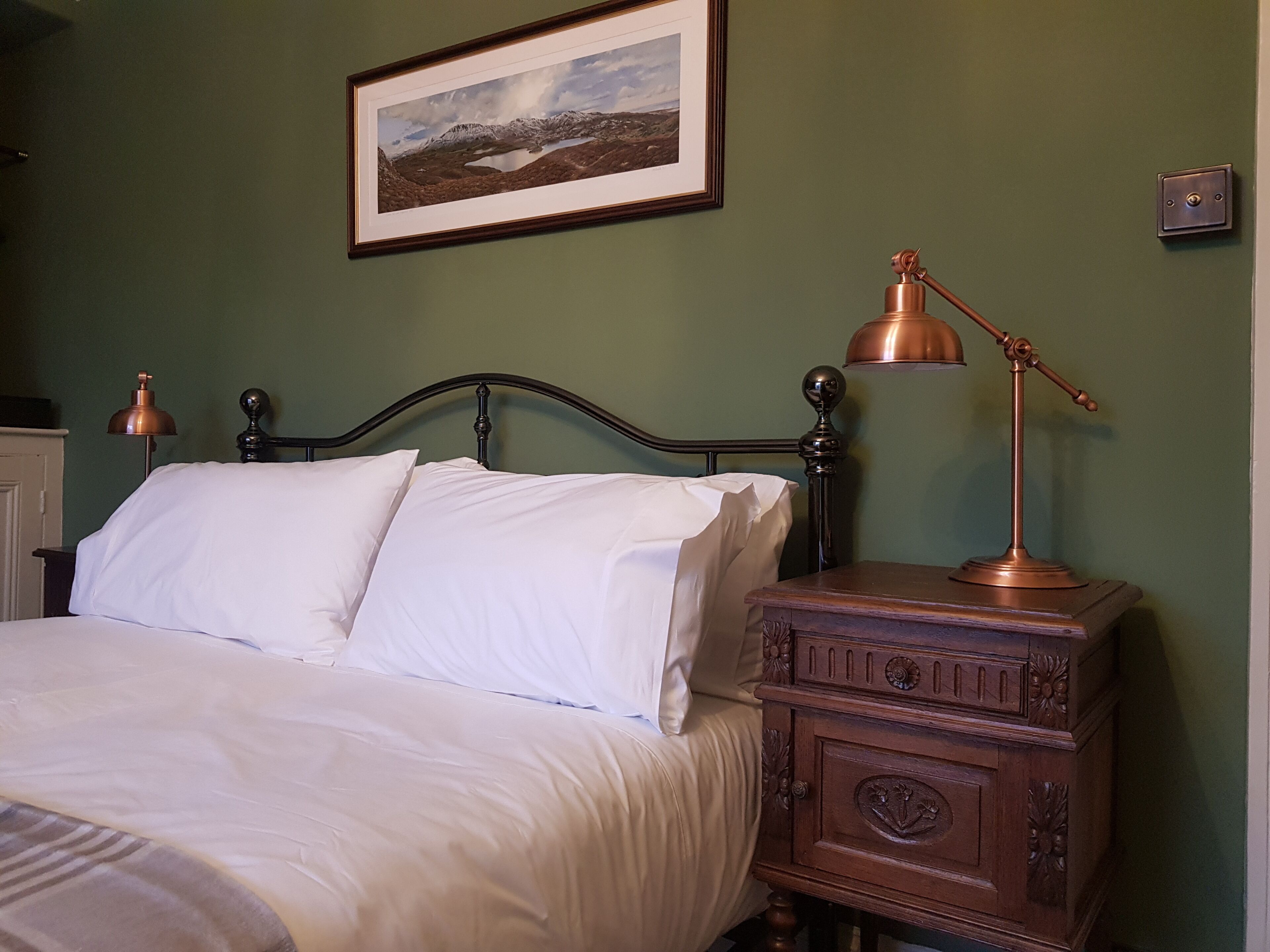 Un lit bien fait, avec du linge de lit blanc, une tête de lit en métal foncé et une table de chevet en bois surmontée d’une lampe en cuivre. Un tableau représentant un paysage, encadré, est accroché au mur au-dessus du lit.