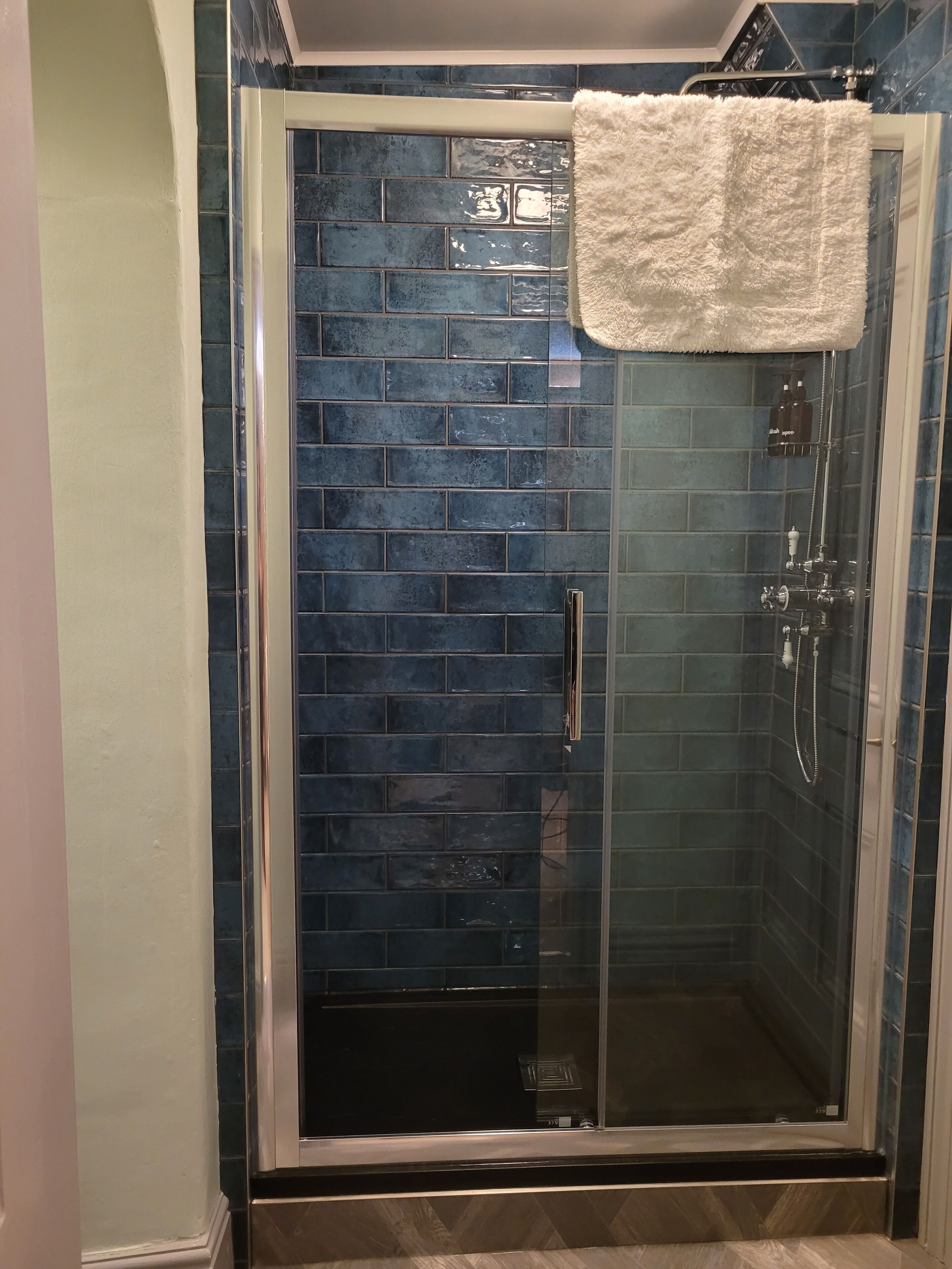 Een glazen douchecabine met een betegelde wand, een witte handdoek hangend aan de deur en een douchekop.
