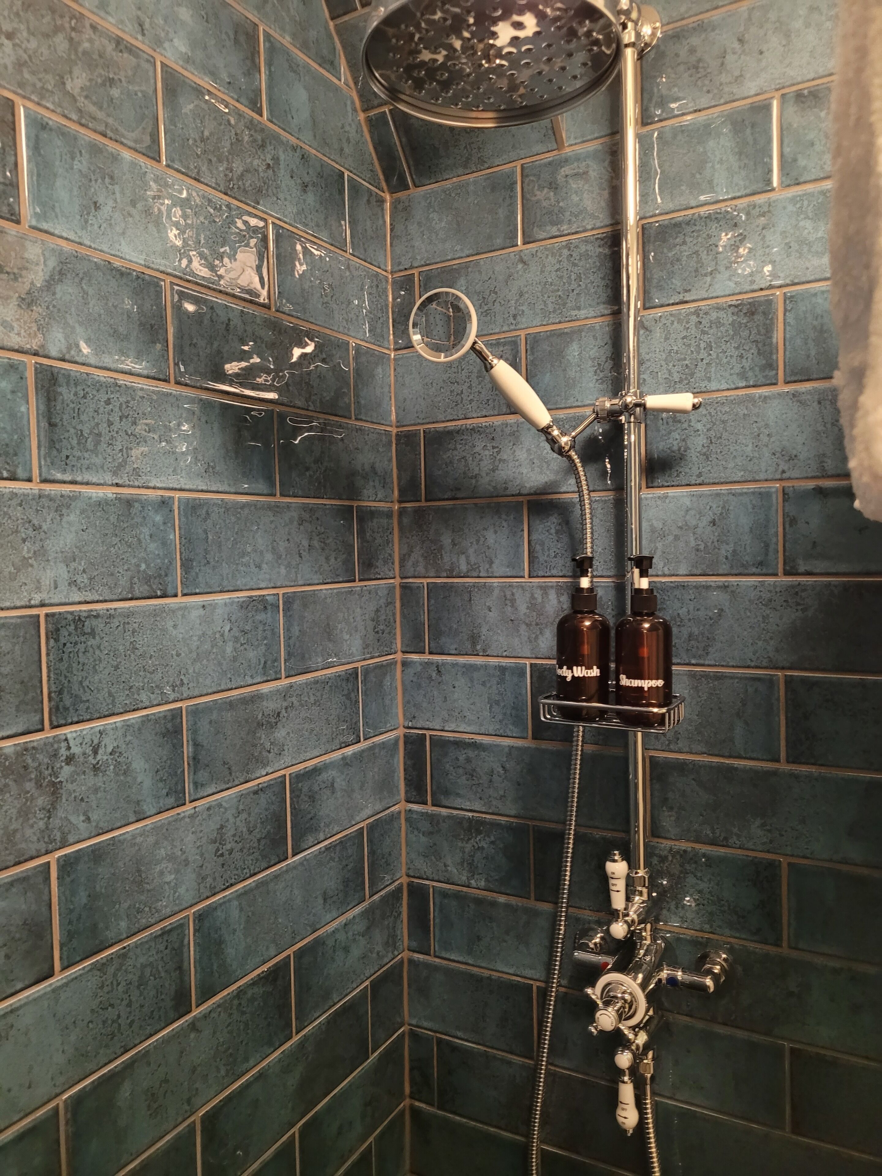 Una ducha con cabezal de ducha de mano y otro cabezal de lluvia empotrado en la pared, ambos con accesorios cromados. Hay dos botellas de champú en un estante adosado a la ducha.