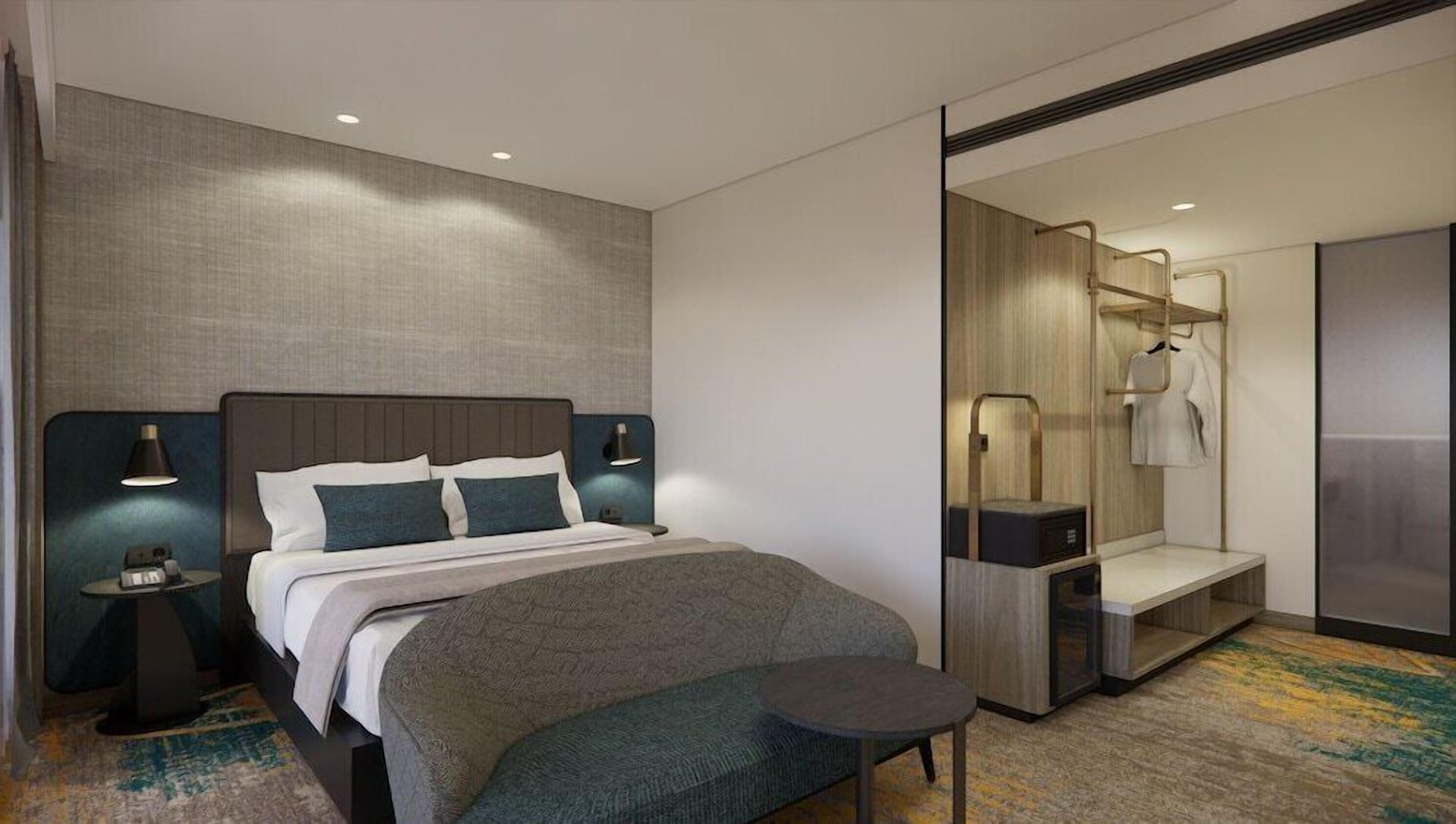 A modern hotel room with a large bed, bedside lamps, a small table, a bench, a walk-in closet, and a mirror.