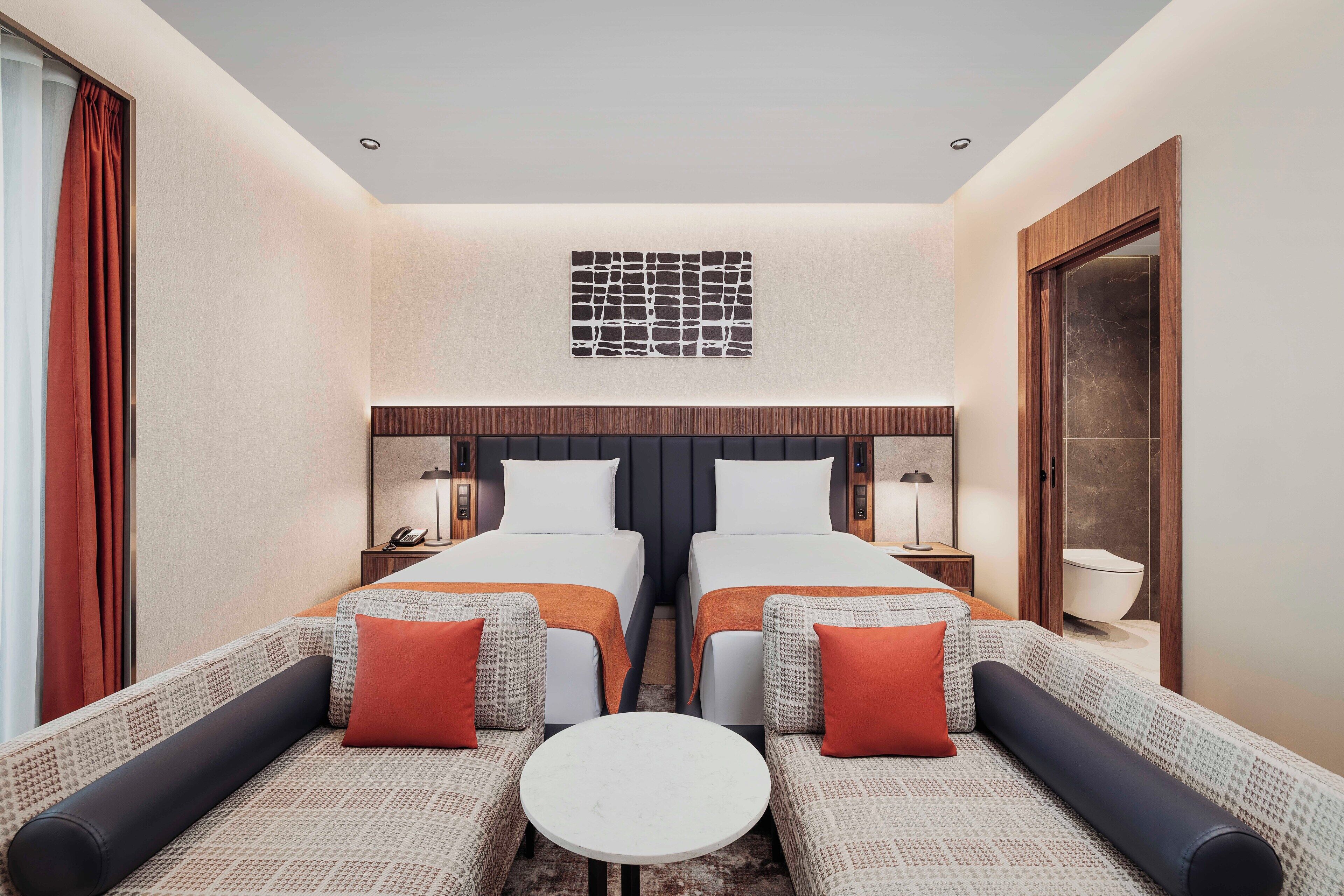 Habitación de hotel con dos camas, un sofá, una mesita y un baño visible a través de una puerta.