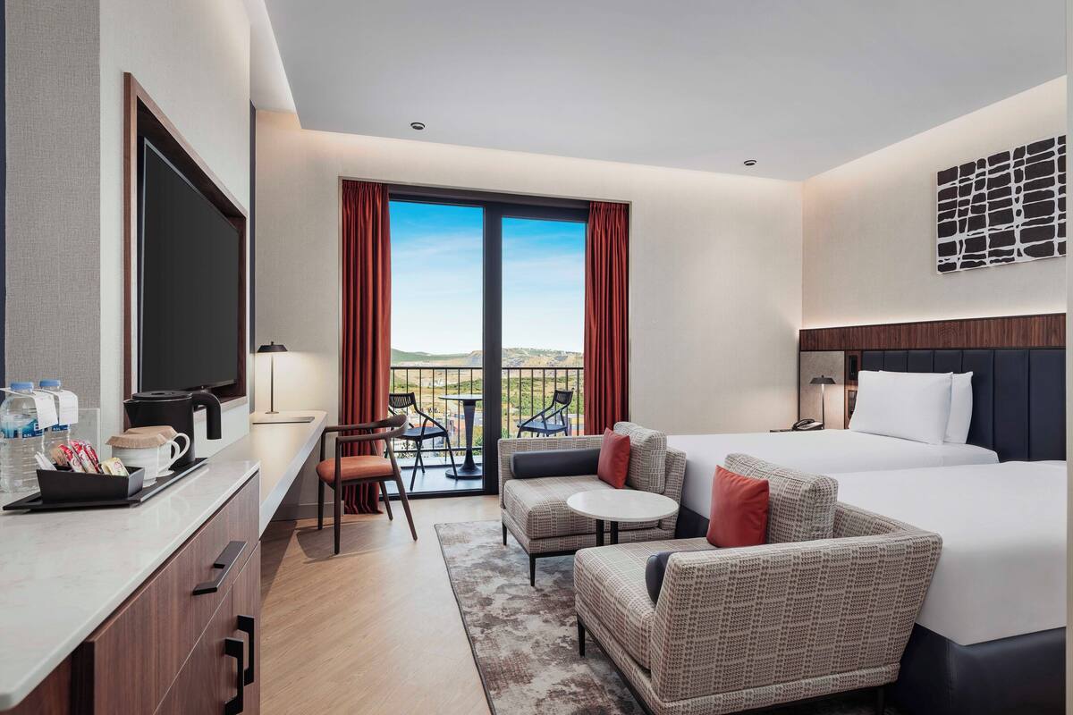 Habitación de hotel con una cama grande, un sofá, un escritorio con televisor y un balcón con vistas.