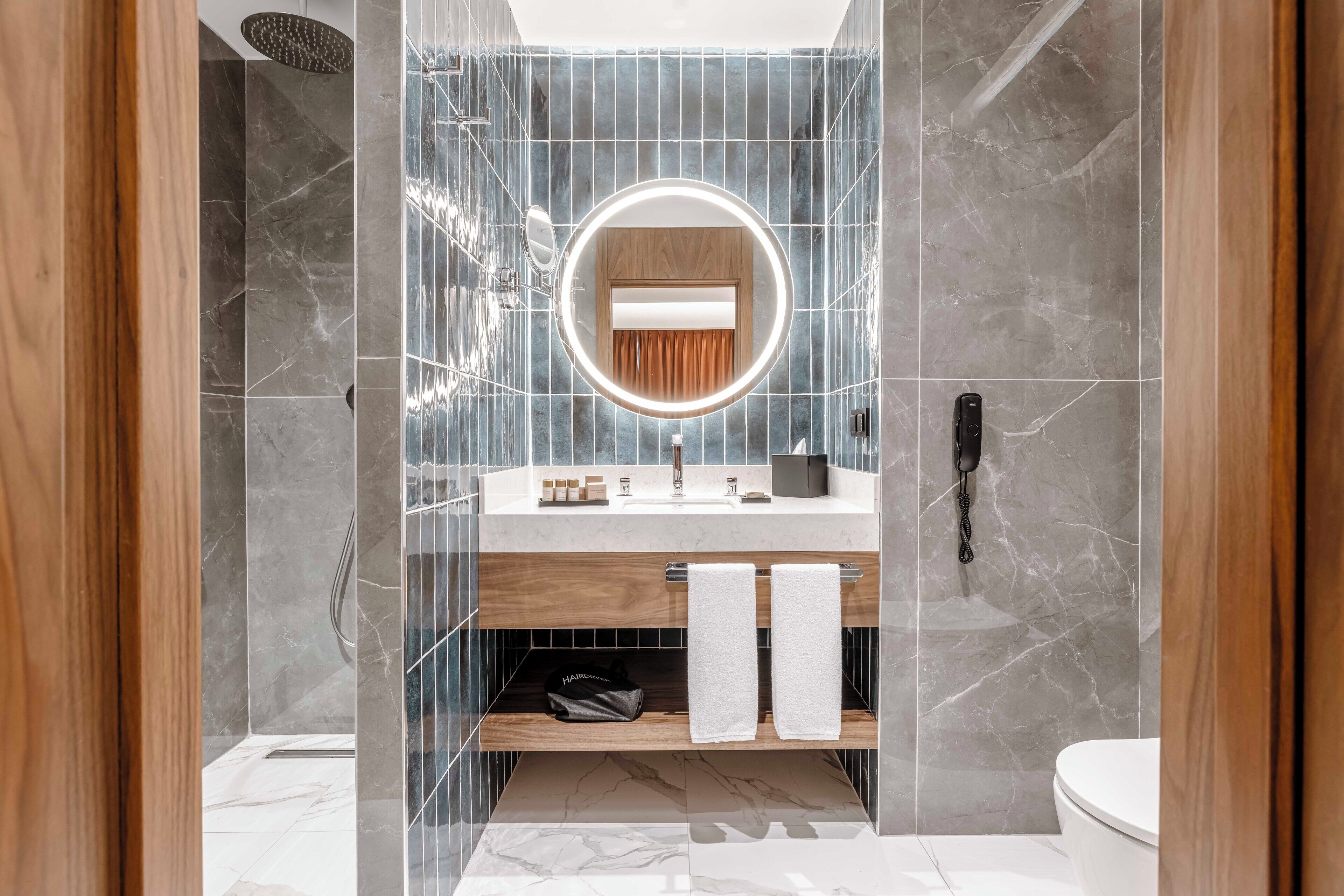 Un baño moderno con ducha revestida de mármol, tocador de madera, espejo redondo con luz e inodoro.