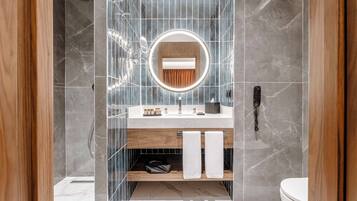 Un baño moderno con ducha revestida de mármol, tocador de madera, espejo redondo con luz e inodoro.