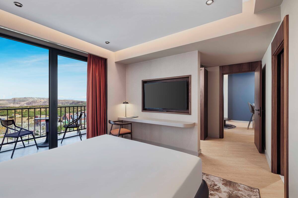 Habitación de hotel con balcón, una cama, televisión montada en la pared y un escritorio con silla.