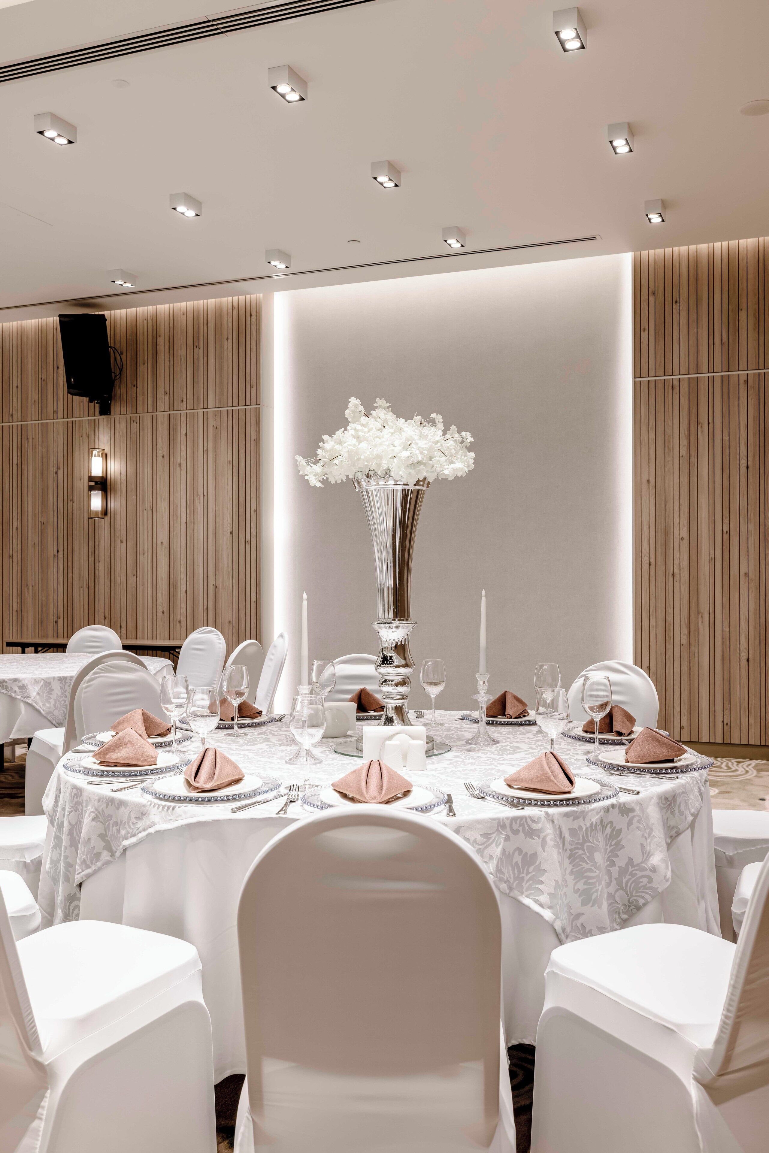 Un comedor formal con mesas redondas, sillas blancas y una elegante vajilla, que incluye un gran jarrón de flores.