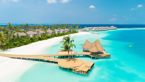 Des bungalows sur pilotis, dotés de toits de chaume, reliés par des passerelles en bois, et offrant une vue imprenable sur une mer turquoise et une plage de sable blanc.