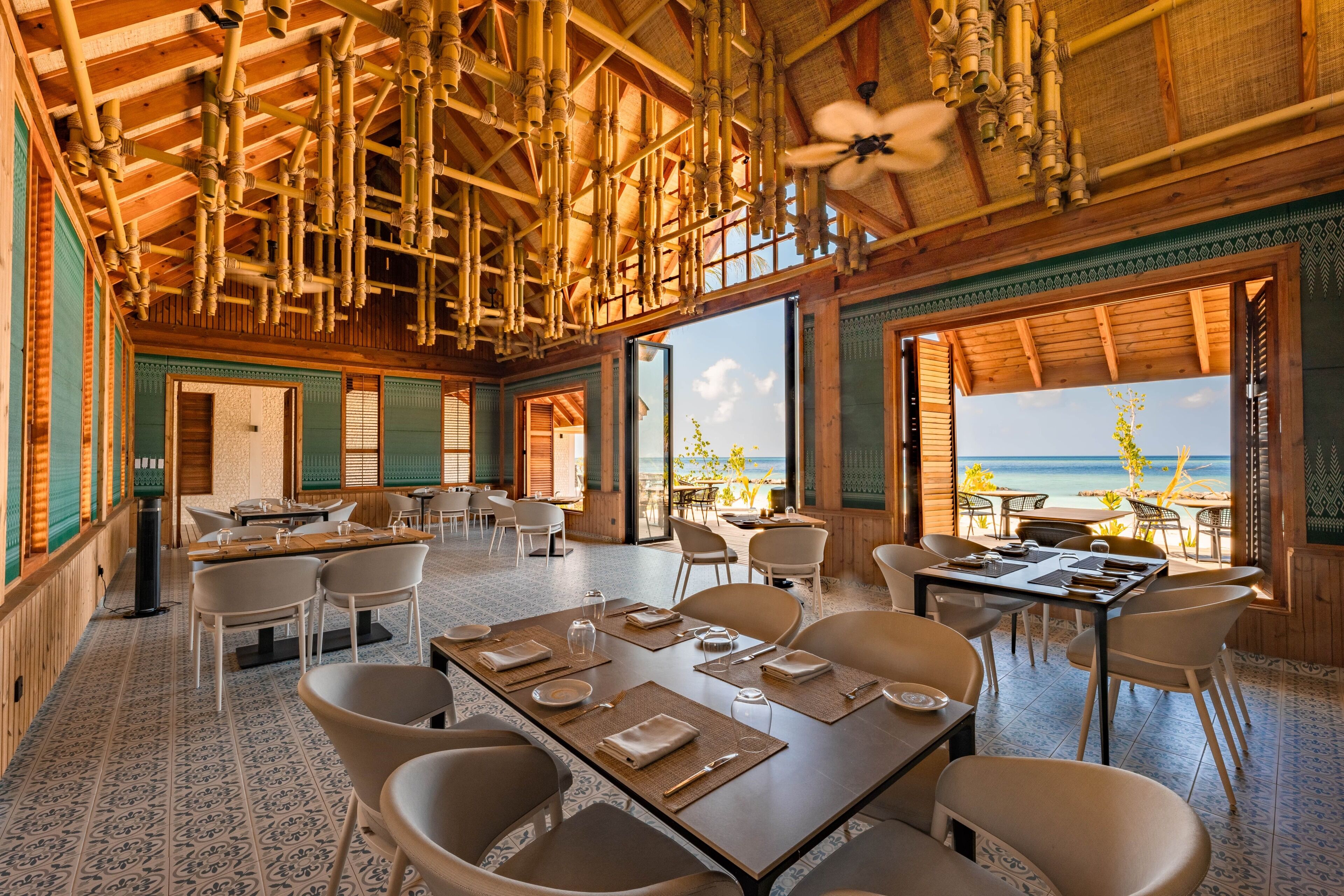 Un comedor espacioso con mesas dispuestas para recibir a los invitados, ventanas amplias con vistas al océano y un techo con detalles de bambú.