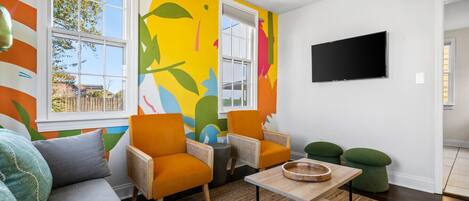 Ein modernes Wohnzimmer mit einem farbenfrohen abstrakten Wandgemälde, zwei orangefarbenen Sesseln, einem grauen Sofa, einem Holztisch und einem flachbildfernseher, der an der Wand montiert ist.