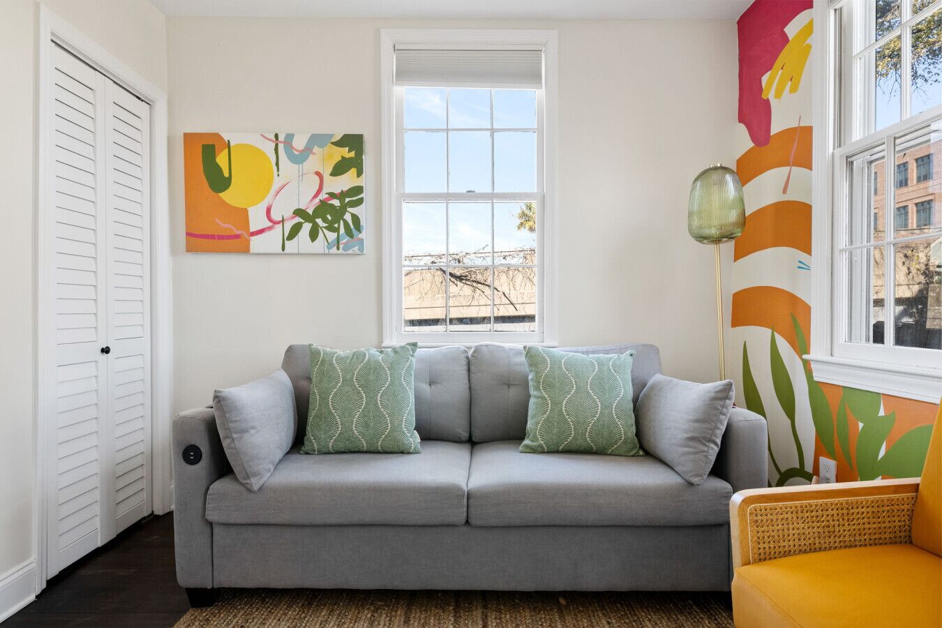 Un salon avec un canapé gris, deux coussins à motifs, une fenêtre donnant sur un beau paysage et un tableau abstrait coloré accroché au mur.