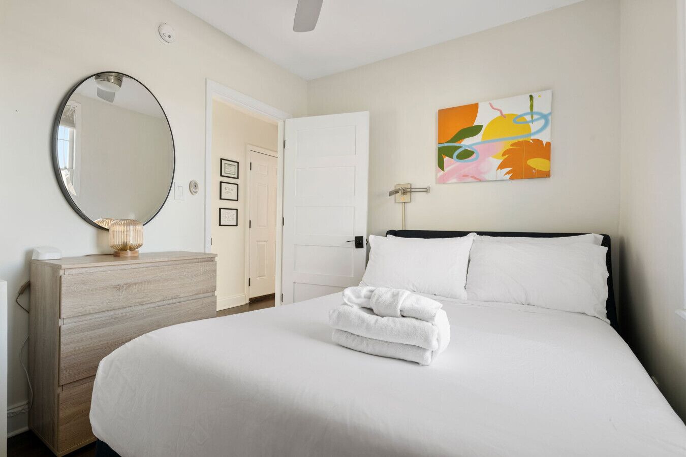 A hotel room with a large bed, a wooden dresser, a round mirror, and a colorful abstract painting on the wall.