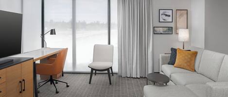 A modern hotel room with a large window, a desk, a chair, a sofa, and a TV.