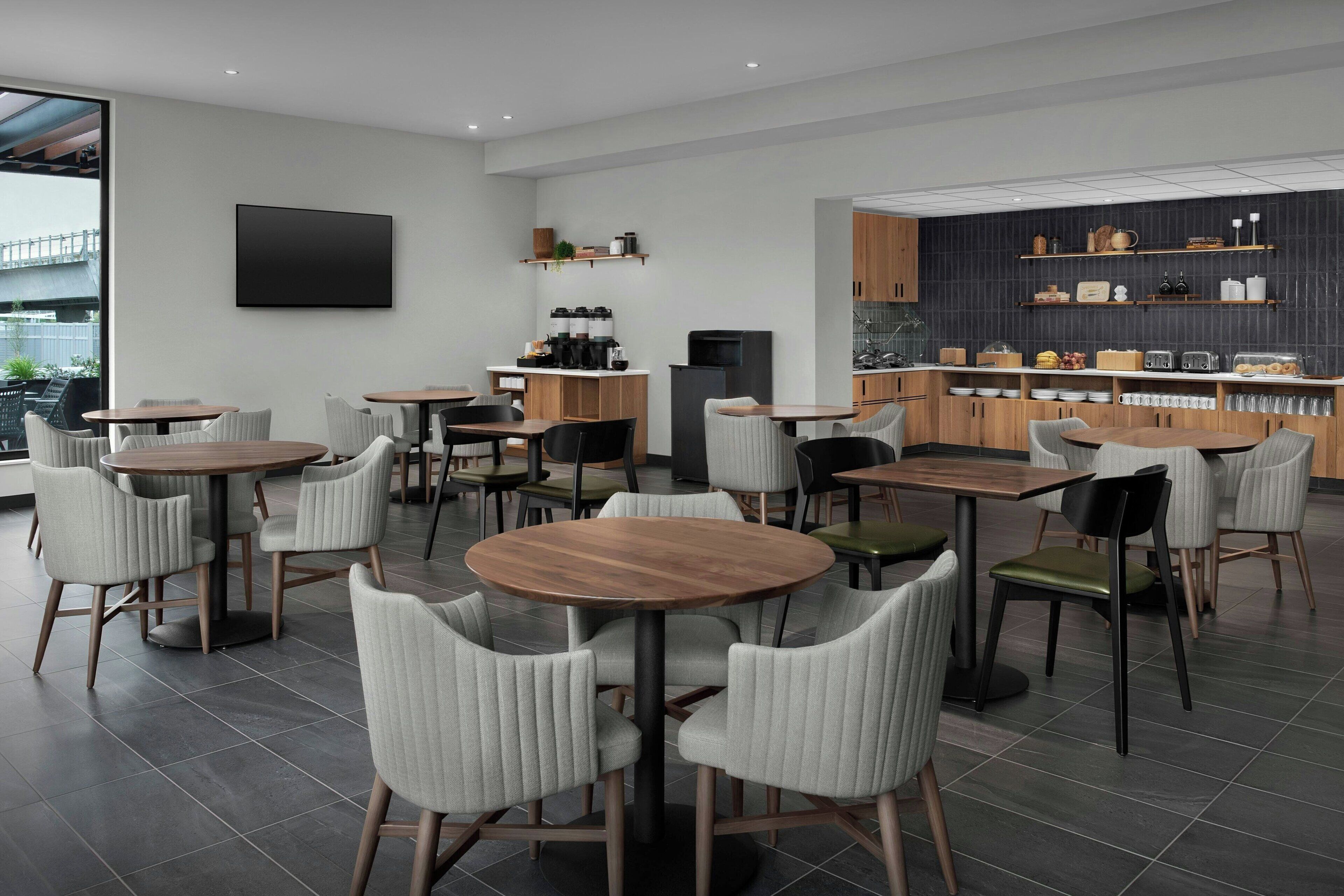 A modern dining area with round tables and chairs, a kitchen with wooden cabinets, and a large flat-screen TV mounted on the wall.