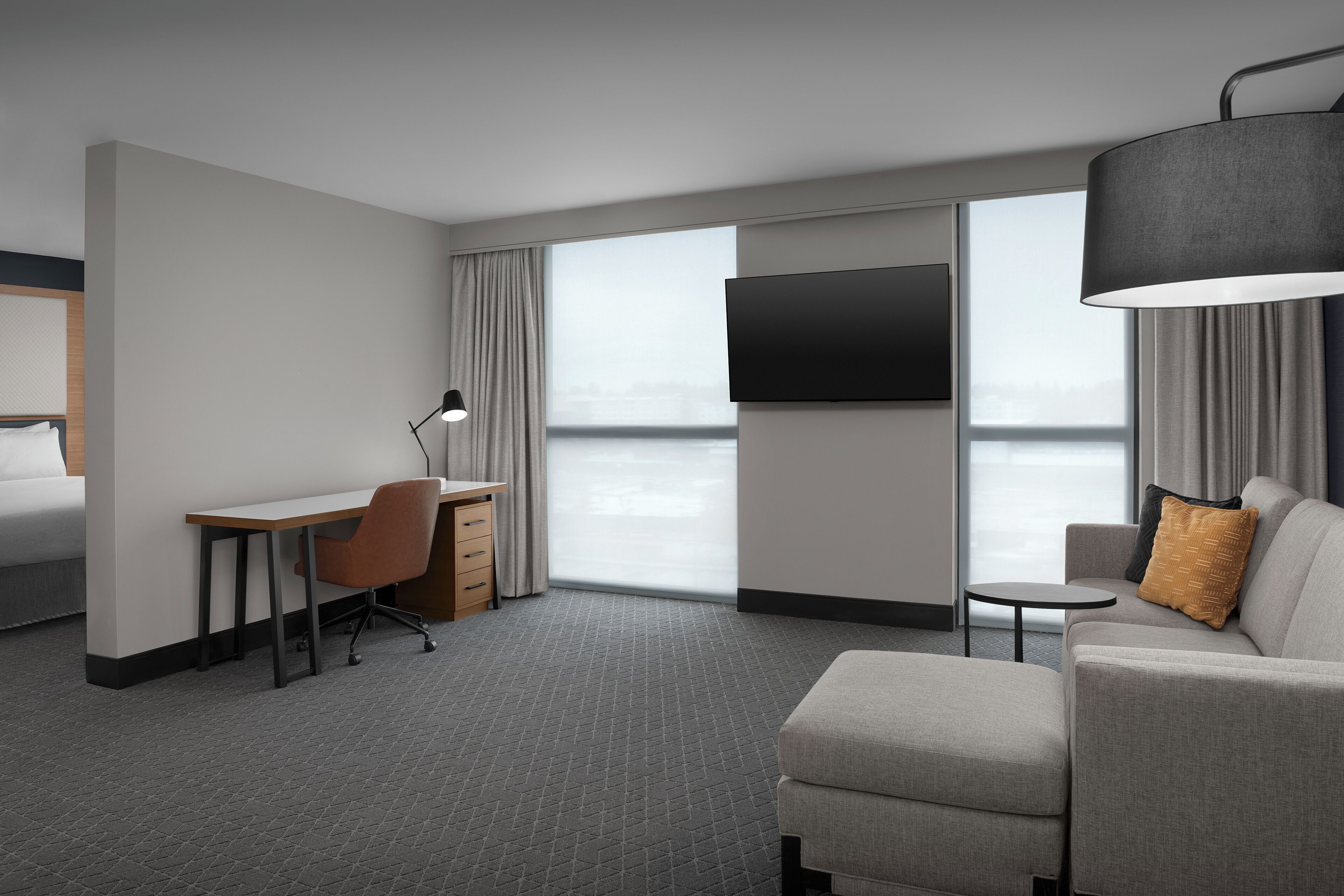 A modern hotel room with a large window, a flat-screen TV, a desk with a chair, a sofa, and a small table.