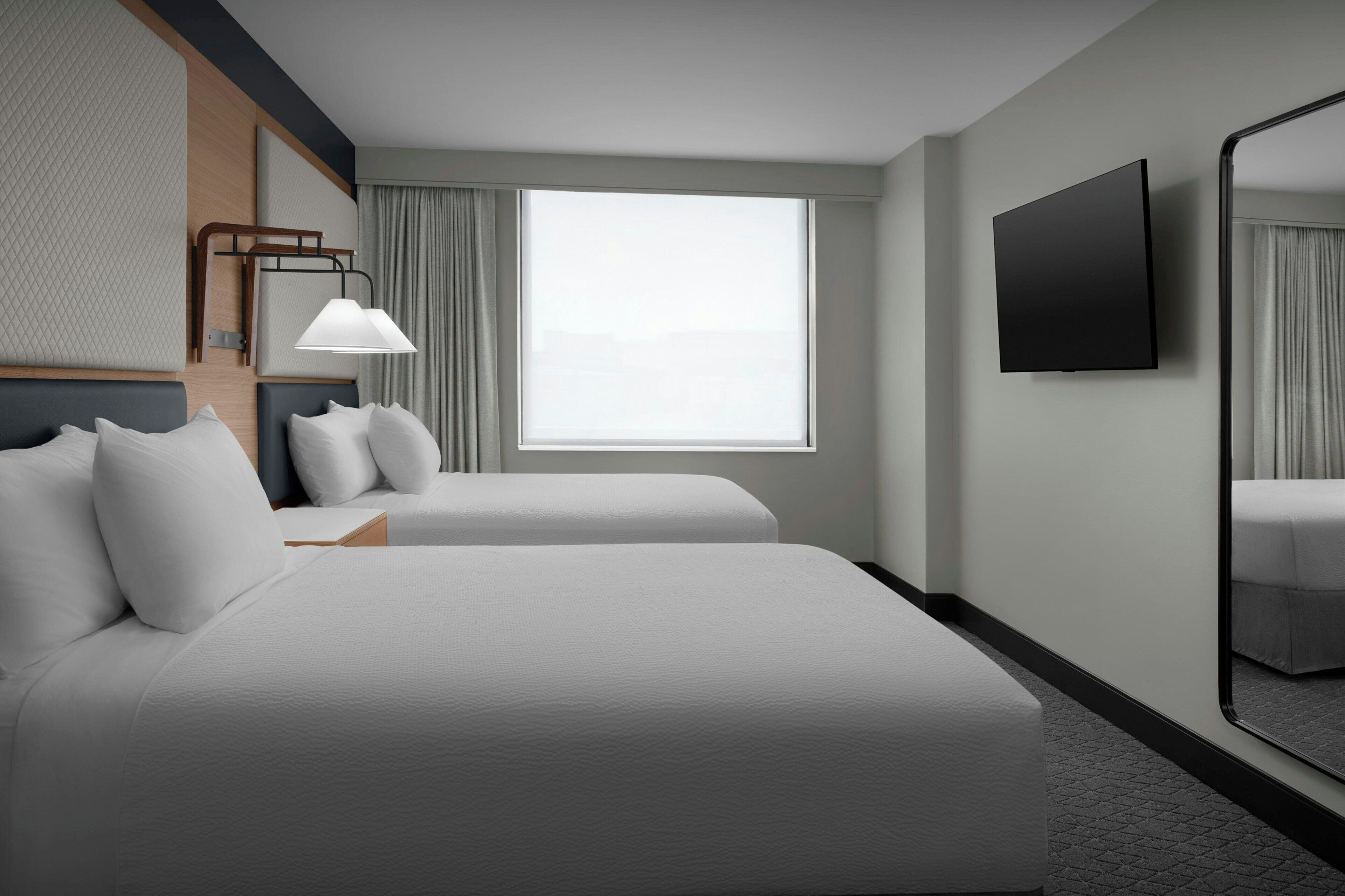 Quarto de hotel com duas camas, uma janela grande, uma TV de tela plana e um espelho.
