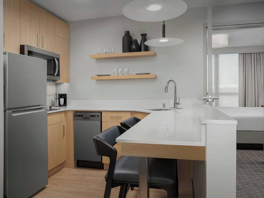 A modern kitchen with a refrigerator, microwave, and dishwasher, wooden cabinets, and a dining area with a white countertop and black chairs.