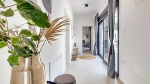 Un couloir moderne agrémenté d’une grande plante en pot, d’un banc rond et offrant une vue sur une chambre avec un lit et un luminaire suspendu.