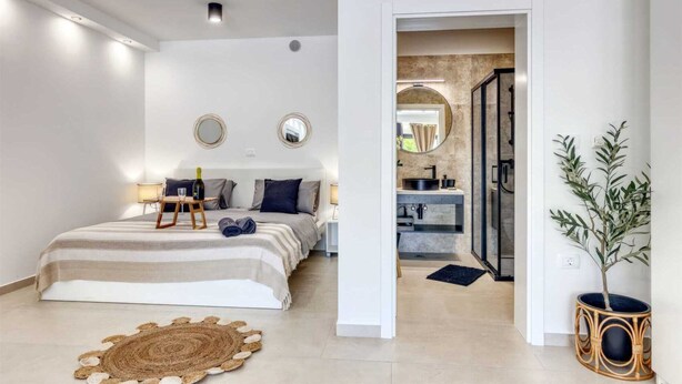 Une chambre moderne avec un grand lit, des tables de chevet et une petite table de nuit. On aperçoit une salle de bain à travers une porte ouverte, avec un lavabo et un miroir.