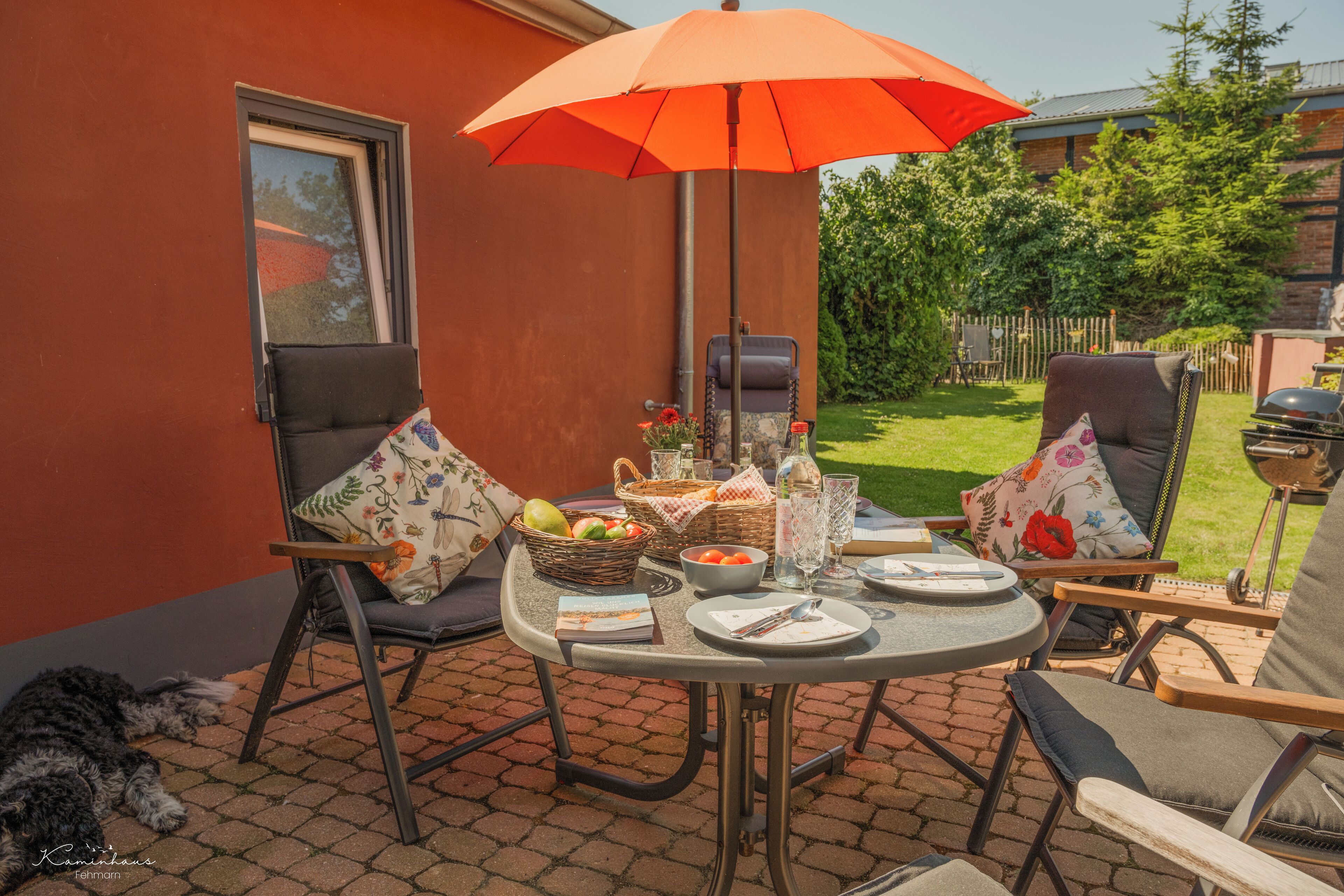 A patio with a table set for two, surrounded by chairs, a basket of fruits, and a grill.