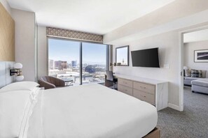 A modern hotel room with a large bed, a desk, a chair, a TV, and a city view.