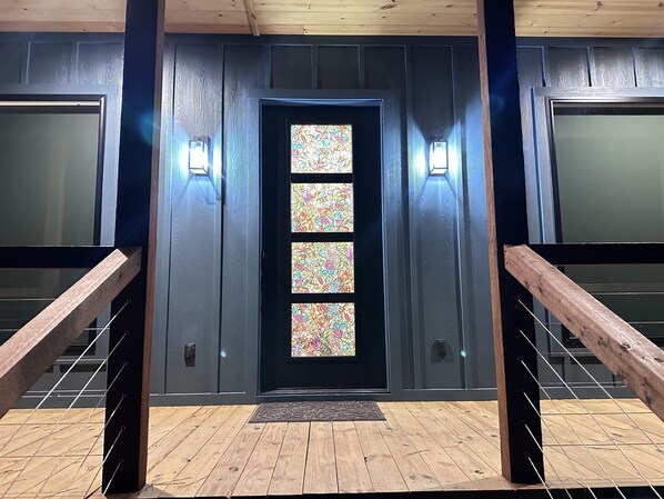 Una entrada moderna con una escalera de madera, una puerta negra con paneles de vidrio emplomado e iluminación en la pared.