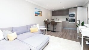 En moderne stue med en grå sofa, et lille hvidt bord og et køkken i baggrunden.