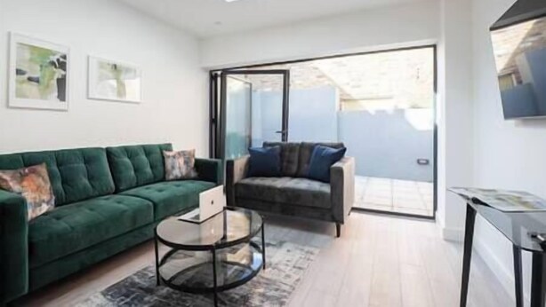 A modern living room with a green sofa, a round glass coffee table, and a grey sofa with blue cushions. There is a sliding glass door leading to a balcony. - PBC | Studio Apt in Richmond (RICH3) (London)