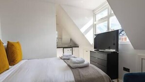 A modern bedroom with a bed, a TV, and a kitchenette in the background.