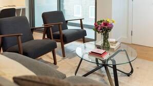 A modern living room with two grey armchairs, a glass coffee table with books and a vase of flowers, and a view of a brick building through a glass door.