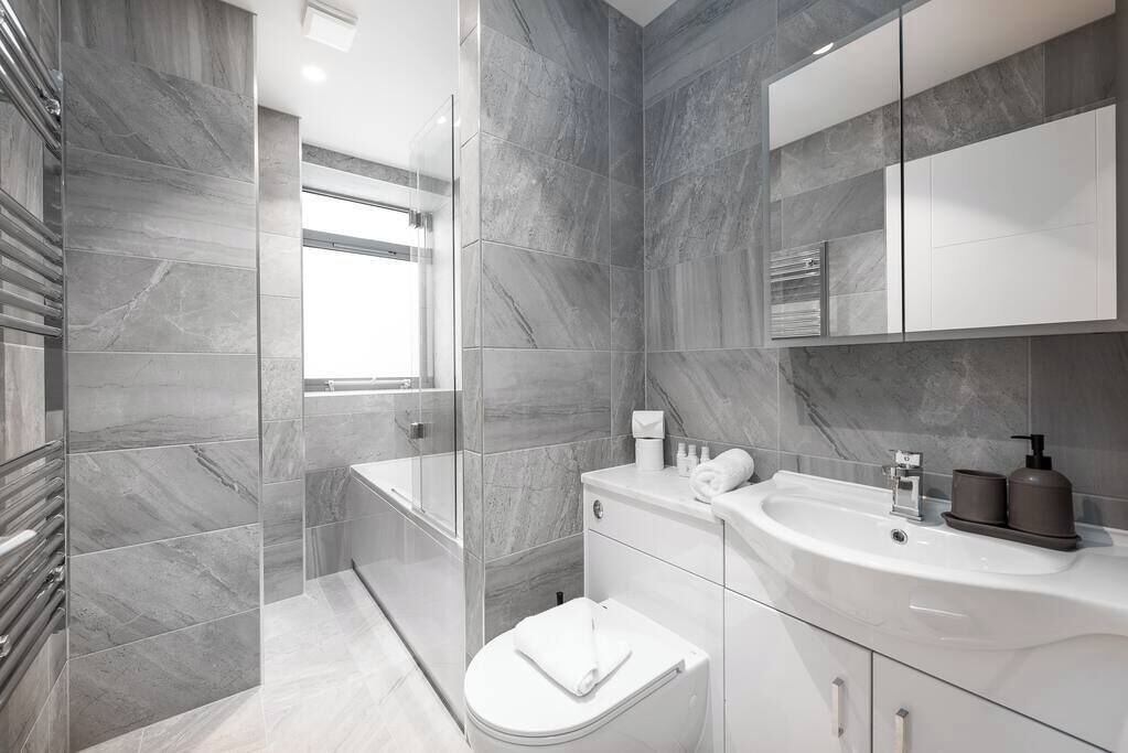A modern bathroom with grey marble tiles, a glass shower enclosure, a white toilet, and a vanity with a sink and mirror.