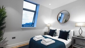 Une chambre d’hôtel moderne avec un grand lit, deux tables de chevet, un miroir rond et une fenêtre donnant sur un immeuble.