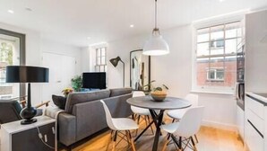 Un salon moderne avec un canapé gris, une table de salle à manger ronde entourée de chaises blanches, une lampe noire et un téléviseur posé sur un meuble.