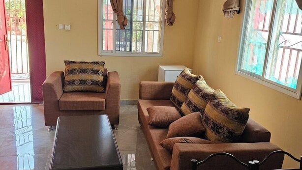 A living room with a sofa, a coffee table, and a dining table. There are curtains on the windows, and a radiator is visible on the wall. - Kaira kunda (Sanyang)