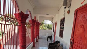 A corridor with a tiled floor, red pillars, and a red door with a geometric pattern. There are plastic chairs along the wall and a white chair near a window. - Kaira kunda (Sanyang)