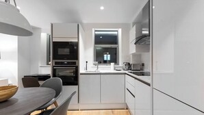A modern kitchen with white cabinetry, a built-in oven, and a window above the sink. - PBC | Duplex Two Bedroom Apartment in Shoreditch (London)