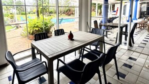 A covered patio area with a table and chairs, large windows, and a view of a pool and garden. - Coral house San Andres (San Andrés)