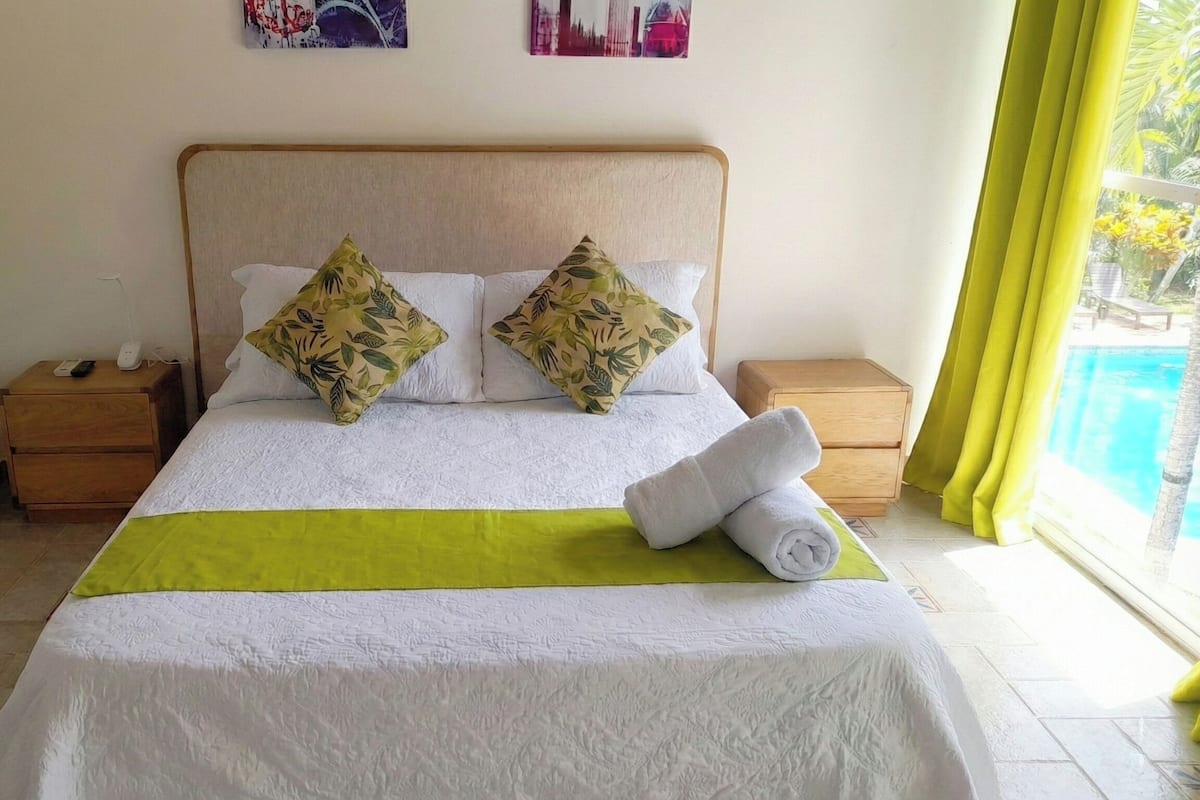 A neatly made bed with a green bedspread, two decorative pillows, and a rolled towel on the side. There are two bedside tables, one with a phone and charger, and a window with yellow curtains.