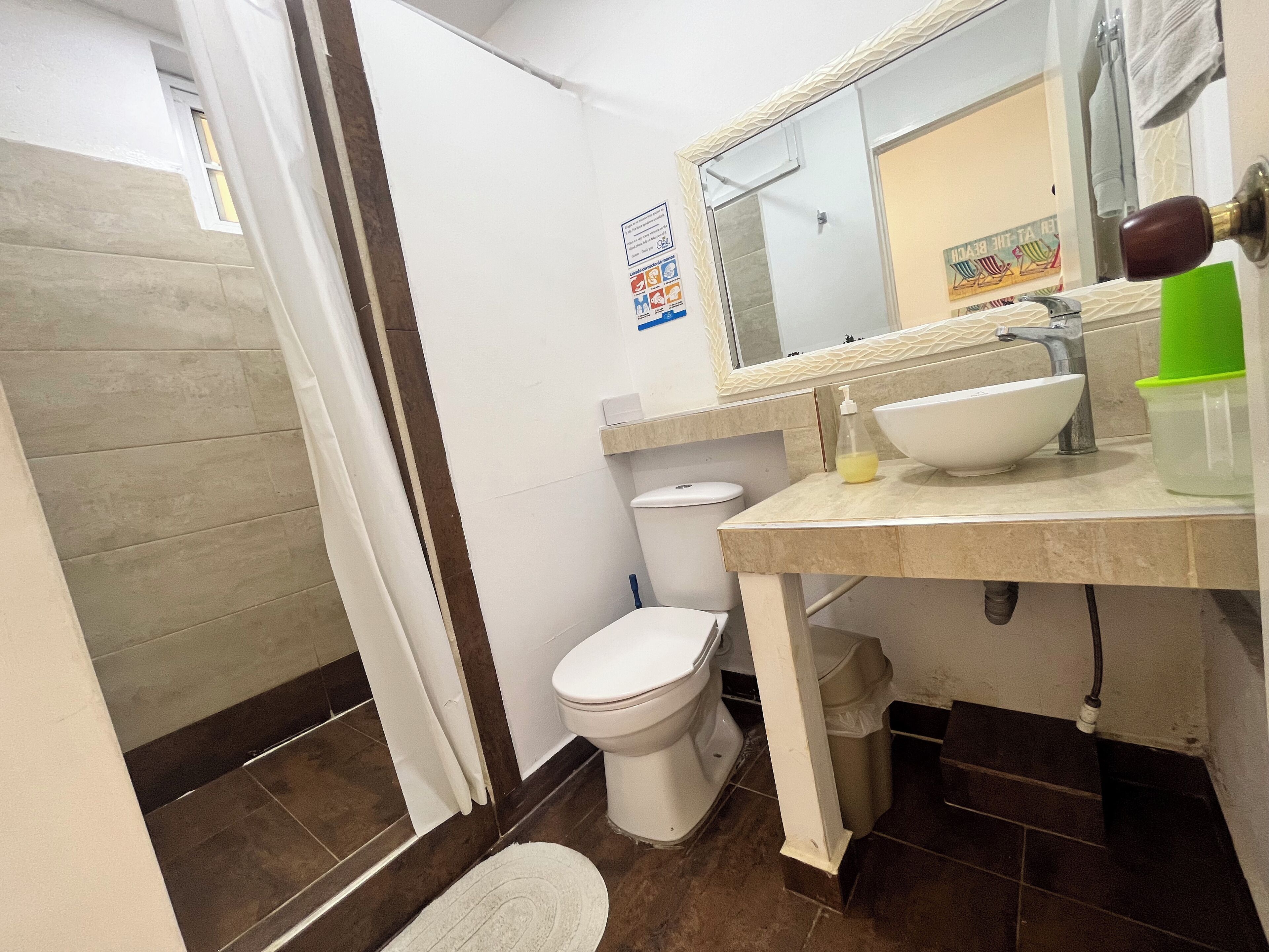 Egy fürdőszoba, amelyben WC, mosdókagyló és tükör található. Van egy zuhanyfüggöny, egy törölközőtartó és egy poszter a falon.