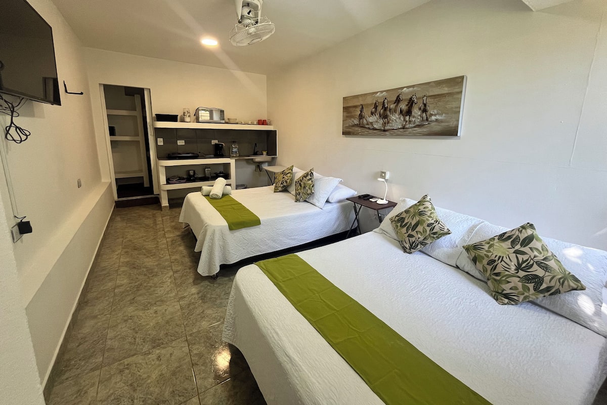 A hotel room with two beds, a painting on the wall, a ceiling fan, and a kitchenette area.