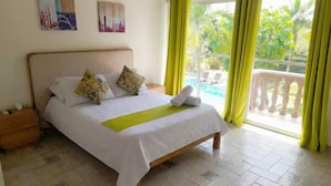 A neatly made bed with a white bedspread and two floral pillows, a wooden nightstand with a lamp, and a view of a pool through large windows with yellow curtains. - Coral house San Andres (San Andrés)