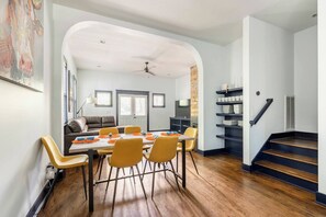 Un comedor con mesa, sillas y sofá. Se ve una escalera con barandilla y una puerta que da a otra habitación.