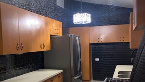 A compact kitchen with wooden cabinets, a stainless steel refrigerator, a built-in oven, and a black countertop. - D’Emerald of the Sea - Unit 5, Mid-Island 2-Bed Modern Apartment near the Beach (Christiansted)
