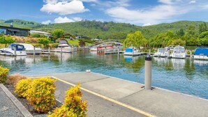 A marina with boats docked, a pedestrian walkway, and a backdrop of hills and a building.
