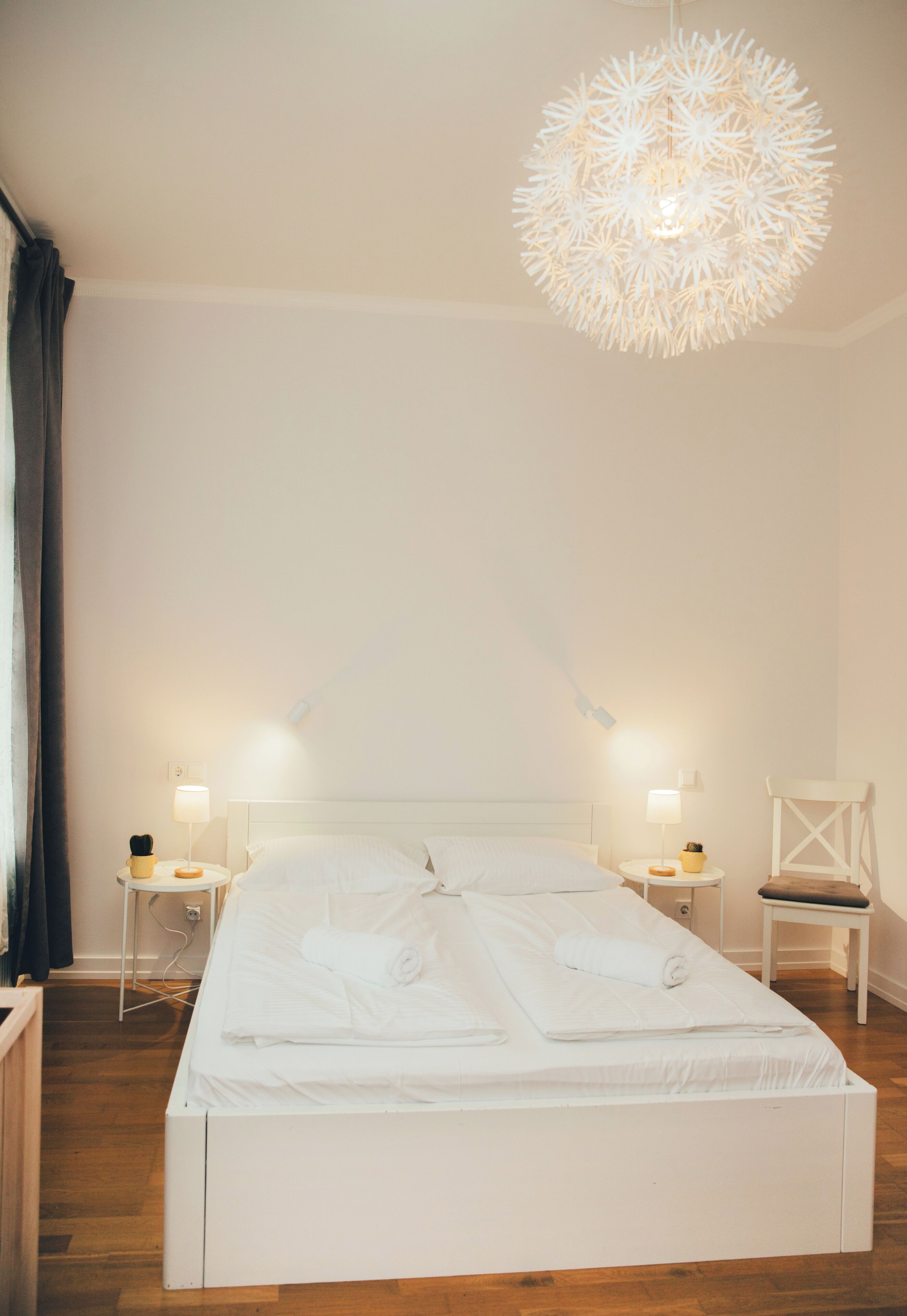 Uma cama bem arrumada com lençóis brancos, duas mesas de cabeceira com luminárias, uma cadeira branca e um lustre esférico único.