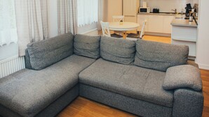 A modern living room with a grey sectional sofa, a dining table with chairs, and a kitchen area in the background. - Beautiful, cozy, family-friendly apartment in the middle of nature. (Sigulda)