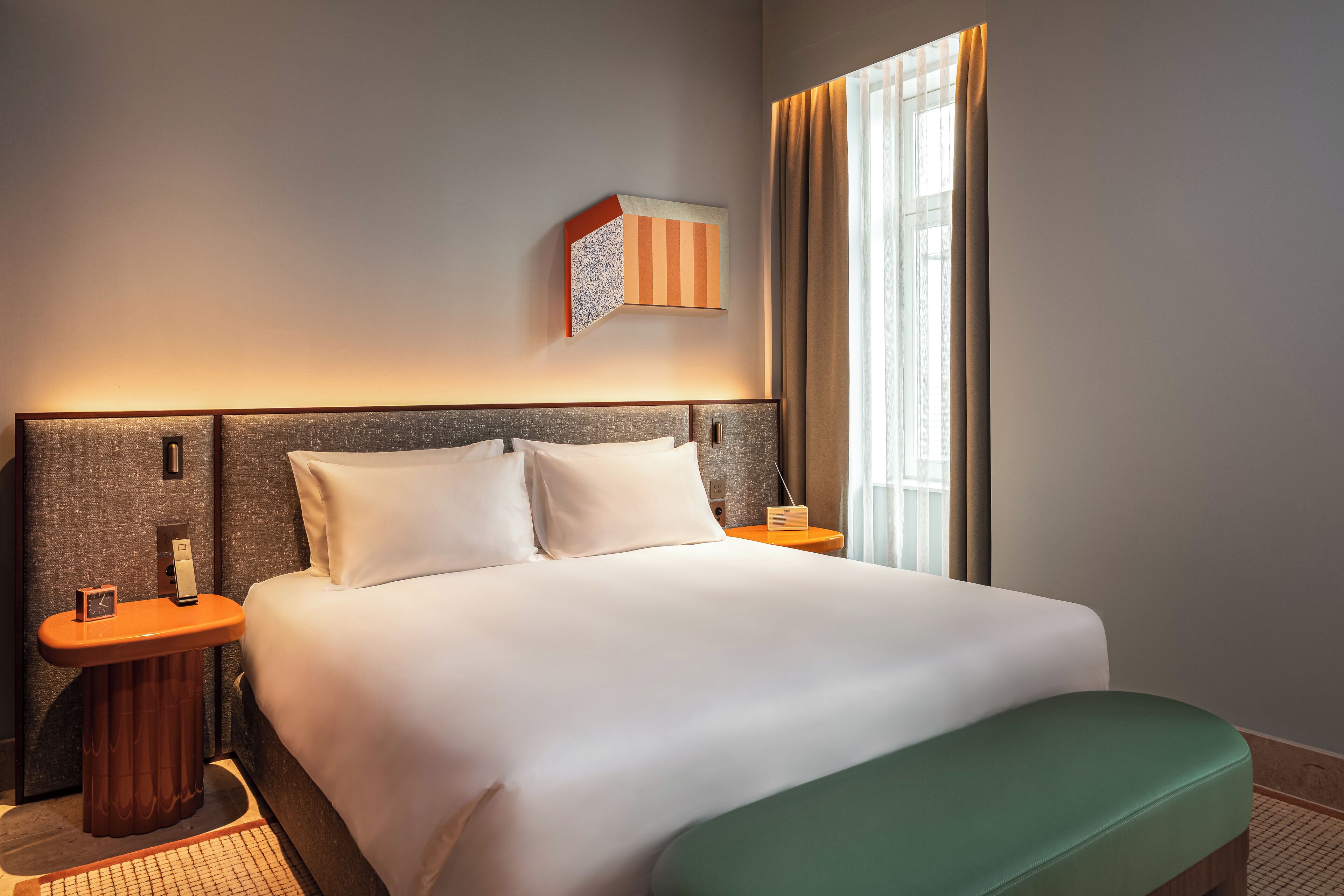 A hotel room with a large bed, a nightstand with a lamp, a wall-mounted artwork, and a window with curtains.